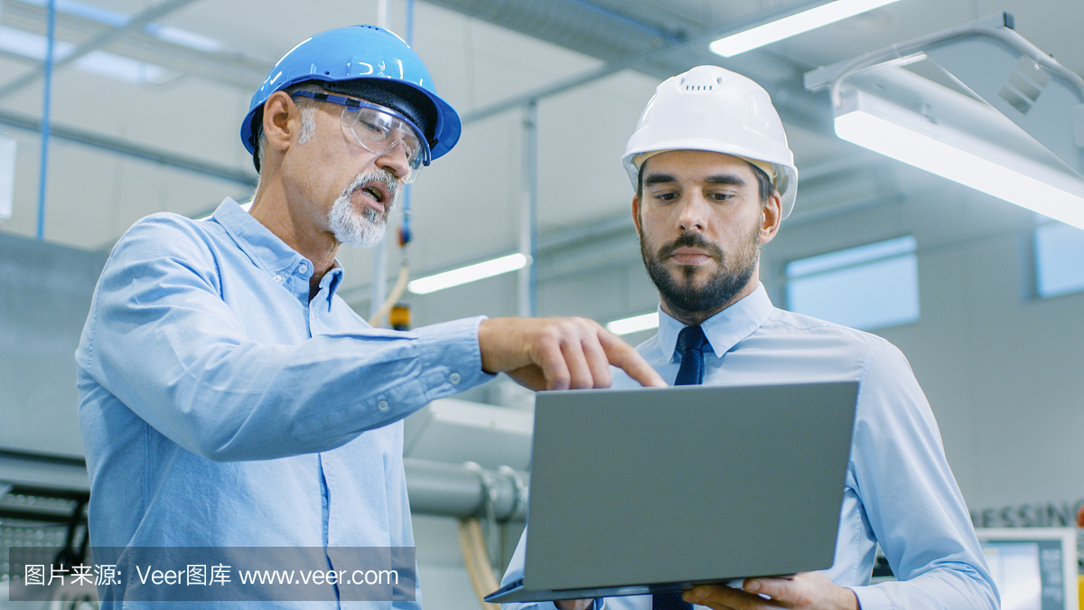 部門主管持有筆記本電腦并與總工程師討論產(chǎn)品細(xì)節(jié)。他們戴著安全帽在現(xiàn)代工廠工作。
