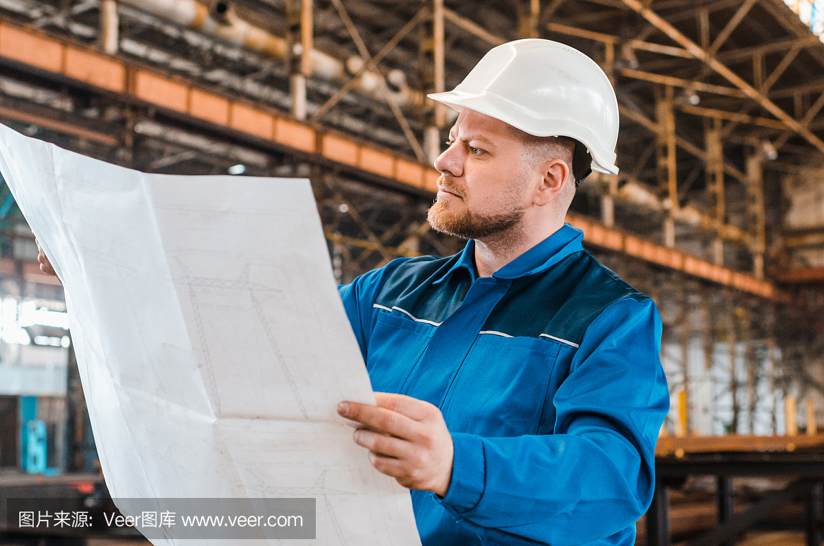 一個(gè)穿著工作服、戴著白色頭盔的工人正在檢查一個(gè)金屬工廠背景上的圖紙。建筑工人身體很好。男工程師
